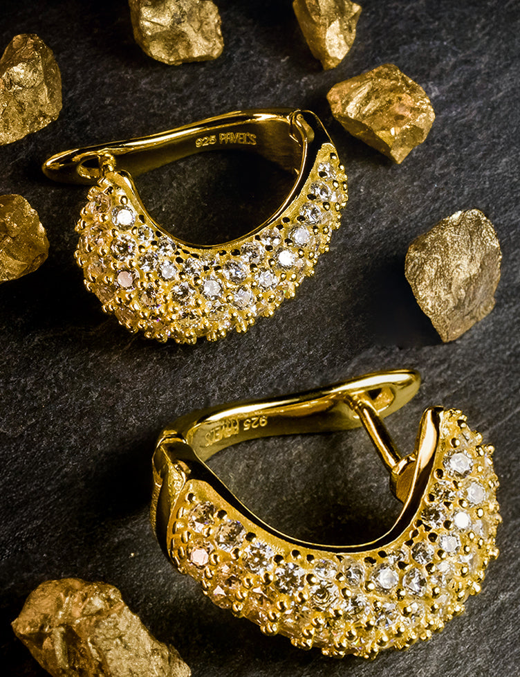 Paar goldene Ohrringe mit funkelnden Diamanten liegt auf dunklem Stein, umgeben von goldenen Nuggets.