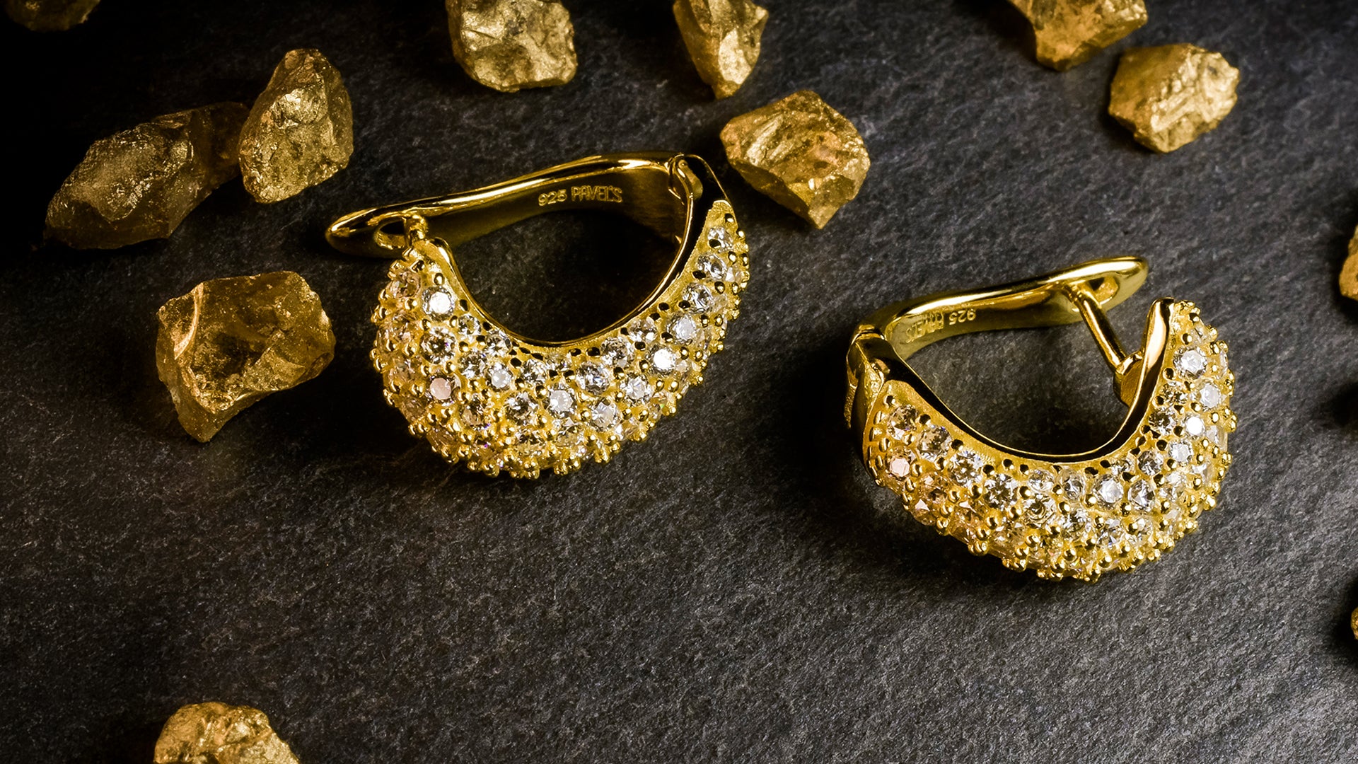 Paar goldene Ohrringe mit funkelnden Diamanten liegt auf dunklem Stein, umgeben von goldenen Nuggets.