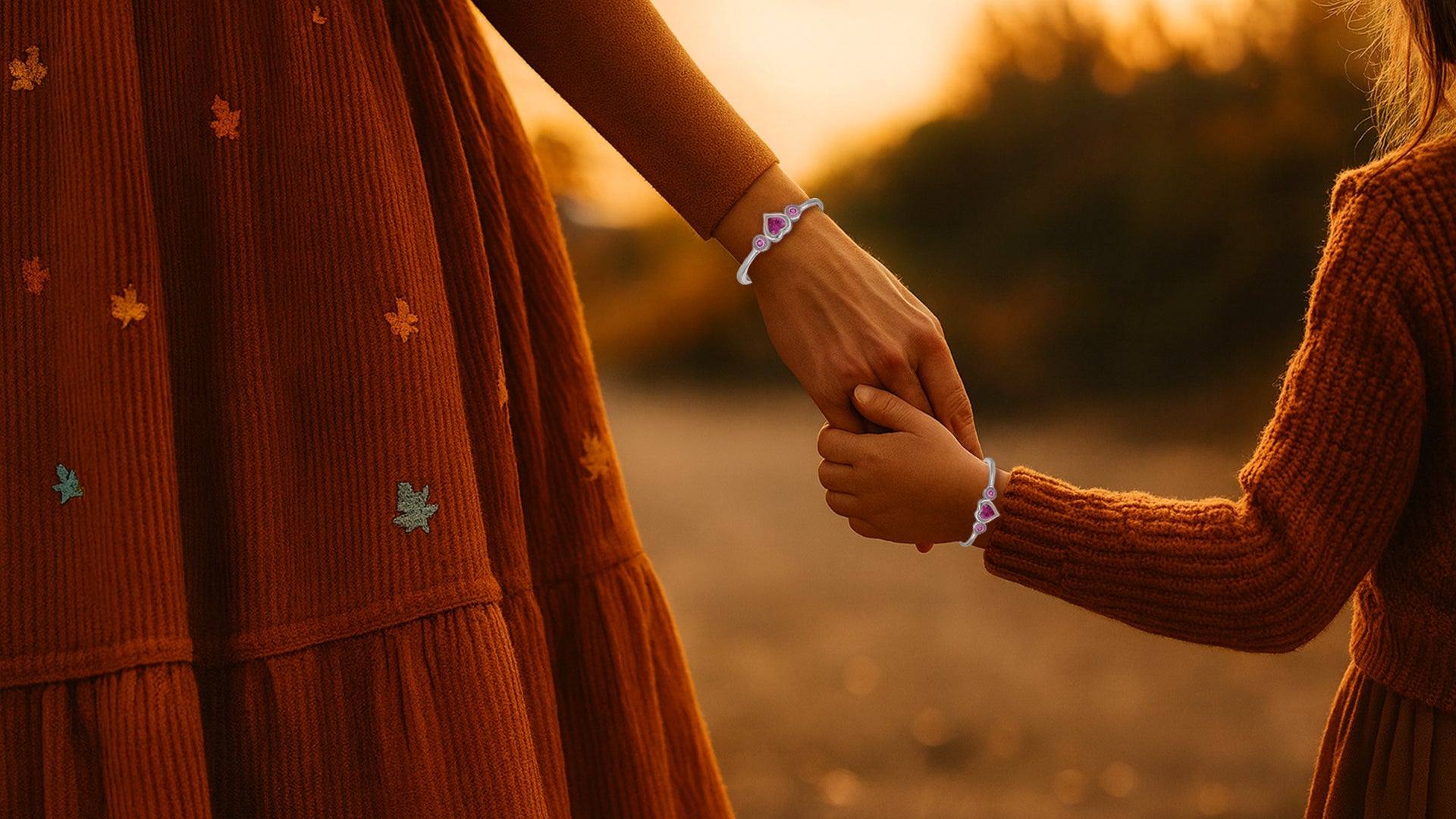 Mutter und Kind halten sich an den Händen und tragen passende Herzarmbänder bei warmem Licht eines herbstlichen Sonnenuntergangs.